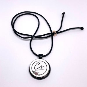 May include: A black cord necklace with a circular pendant. The pendant has a white background with the letters "Cs" in a cursive font, surrounded by a floral design and the text "@COLLECTIONSAGE". The necklace has a gold-colored bead.