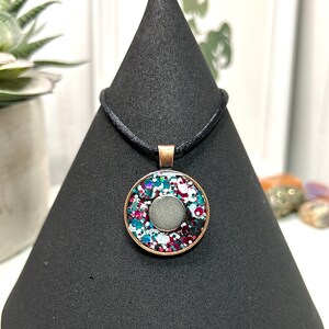 May include: A round pendant necklace with a copper-colored frame. The pendant features a central gray stone surrounded by colorful glitter in shades of teal, red, and white. The necklace has a black cord.