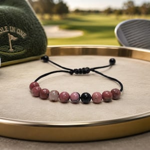 May include: A beaded bracelet featuring a series of round pink and black stones strung on a thin black cord. The bracelet includes an adjustable sliding knot closure with two small black beads at the ends of the cord, creating a simple design.