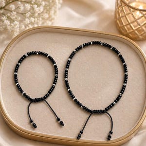 May include: Two black beaded bracelets with silver detailing and adjustable black cords are displayed on a gold-rimmed tray. The bracelets are of different sizes, suggesting a matching set. The background is a soft, neutral colour.