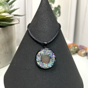 May include: A round pendant necklace with a black cord. The pendant has a black frame with a silver and iridescent center. The necklace is displayed on a black cone-shaped stand.