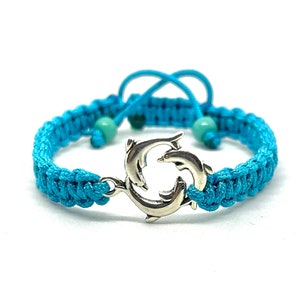Könnte beinhalten: Ein geflochtenes Armband in Türkisblau mit einem silbernen Delfin-Anhänger. Das Armband hat einen verschiebbaren Knotenverschluss und ist mit kleinen türkisfarbenen Perlen verziert.