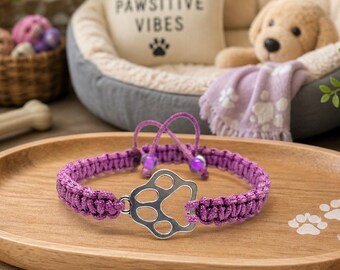 May include: A purple macrame dog collar with a silver paw print charm. The collar is displayed on a wooden tray with paw print accents. Other pet accessories are in the background, including a dog bed and a stuffed animal.