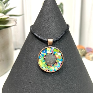 May include: A round pendant necklace with a copper-colored frame. The pendant features a central gray circle surrounded by colorful glitter in shades of blue, green, and pink. The necklace has a black cord.