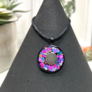 May include: A black pendant necklace with a circular design. The pendant features a black frame filled with colorful glitter in shades of pink, blue, and yellow. A silver-colored circle is in the center. The necklace cord is black.