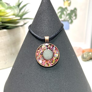 May include: A round pendant necklace with a copper-colored frame. The pendant features a central gray stone surrounded by colorful glitter. The necklace has a black cord.