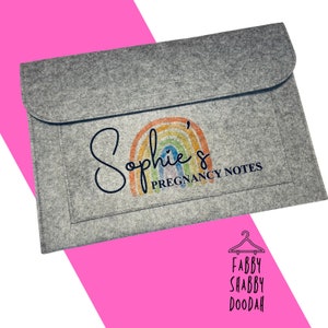 May include: A grey felt document holder with a flap. It has a rainbow design and the text "Sophie's Pregnancy Notes". The bottom right corner has the text "Fabby Shabby Doodah" with a hanger graphic.
