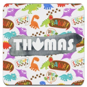 Puede incluir: Un posavasos cuadrado con un fondo blanco con coloridos dinosaurios de dibujos animados, huellas y bocadillos con frases como "RAWWR!" y "STOMP!". El nombre "THOMAS" se muestra prominentemente en letras blancas sobre una forma gris de estilo acuarela.