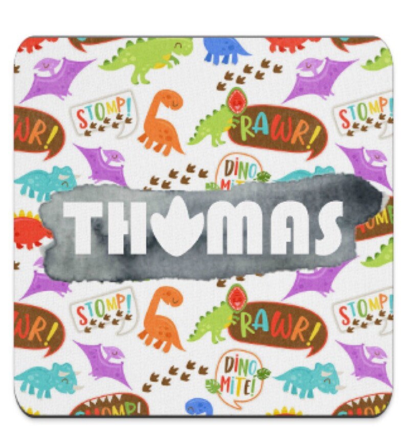 Puede incluir: Un posavasos cuadrado con un fondo blanco con coloridos dinosaurios de dibujos animados, huellas y bocadillos con frases como "RAWWR!" y "STOMP!". El nombre "THOMAS" se muestra prominentemente en letras blancas sobre una forma gris de estilo acuarela.