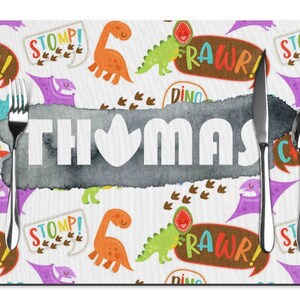 Puede incluir: Un mantel individual personalizado con tem&aacute;tica de dinosaurios. El mantel individual presenta un fondo blanco con ilustraciones de dinosaurios coloridos, bocadillos de texto y el nombre "THOMAS" en letras blancas grandes. Un tenedor, un cuchillo y una cuchara est&aacute;n colocados.