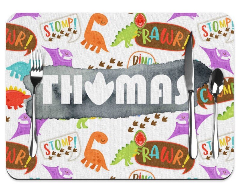Puede incluir: Un mantel individual personalizado con tem&aacute;tica de dinosaurios. El mantel individual presenta un fondo blanco con ilustraciones de dinosaurios coloridos, bocadillos de texto y el nombre "THOMAS" en letras blancas grandes. Un tenedor, un cuchillo y una cuchara est&aacute;n colocados.