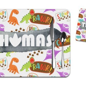Puede incluir: Un juego de manteles individuales y posavasos blancos con tem&aacute;tica de dinosaurios. El mantel individual presenta el nombre "THOMAS" en letras blancas, con un fondo gris estilo acuarela. Dinosaurios coloridos, bocadillos con texto "RAWr!" y "STOMP!" y huellas de dinosaurios decoran el conjunto.