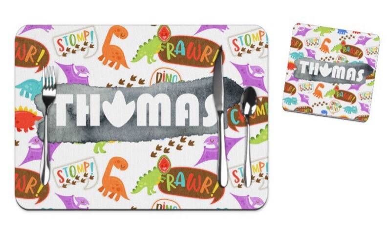 Puede incluir: Un juego de manteles individuales y posavasos blancos con tem&aacute;tica de dinosaurios. El mantel individual presenta el nombre "THOMAS" en letras blancas, con un fondo gris estilo acuarela. Dinosaurios coloridos, bocadillos con texto "RAWr!" y "STOMP!" y huellas de dinosaurios decoran el conjunto.