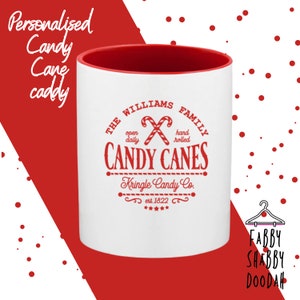 May include: A white ceramic candy cane caddy with a red interior. The caddy features a red graphic with the text "The Williams Family Candy Canes, Kringle Candy Co. est. 1822". The words "Personalised Candy Cane caddy" are in the upper left corner.
