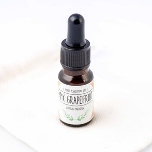 Puede incluir: Una pequeña botella de vidrio ámbar con un gotero negro. La botella está etiquetada con "Pink Grapefruit Pure Essential Oil" y "Citrus Paradisi".