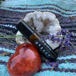 Puede incluir: Un frasco de vidrio marrón con tapa negra y una etiqueta que dice "Achy Breaky". El frasco está sobre una manta de rayas de colores con una geoda de cristal blanco y una piedra roja en forma de corazón.