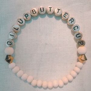 Pode incluir: Um bracelete de contas de madeira brancas com contas de letras douradas que soletram "OLUP BUTTERCUP". O bracelete tem dois pingentes dourados em forma de pirâmide.