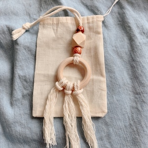 Może przedstawiać: Drewniany pierścień z ozdobnym frędzlem, wiszący na naturalnym bawełnianym woreczku ze sznurkiem. Pierścień ozdobiony jest drewnianymi koralikami w odcieniach brązu i czerwieni.