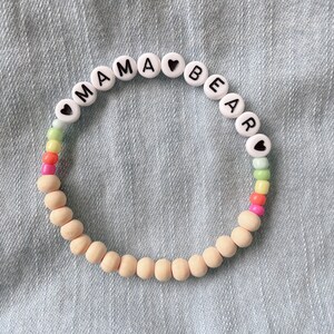 Puede incluir: Un brazalete de cuentas blancas con las palabras "MAMA BEAR" y pequeños corazones entre cada letra. Las cuentas están enhebradas en un cordón beige y tienen cuentas de colores arcoíris intercaladas.