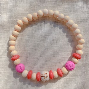 May include: A wooden beaded bracelet with pink and red beads. The bracelet has a wooden bead with a cross design in the center.