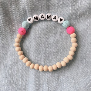 May include: A wooden bead bracelet with the word "MAMA" spelled out in white beads with black lettering. The bracelet also has pink and blue lava beads.