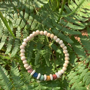 Op de afbeelding: Een witte kralen armband met kleurrijke lavastenen. De armband ligt op een bed van groene varens.