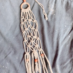 May include: Cream macrame plant hanger with a wooden ring and decorative beads. The plant holder is made of woven cotton cord and features a geometric net design. The top has a wooden bead and a loop for hanging.