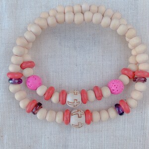May include: Two wooden bead bracelets with pink, red and white beads. One bracelet has a cross charm.