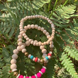 May include: Three beaded bracelets with natural wood beads, gold-colored accents, and colorful accent beads in turquoise, red, and pink. The bracelets are displayed on a bed of green fern leaves.