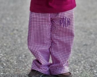 May include: A pair of pink and white gingham pants featuring a purple embroidered monogram with the letters PMR on the upper left thigh, paired with a solid dark pink short-sleeved shirt and brown slip-on shoes.