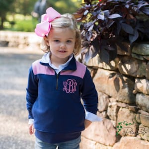 May include: A young child wearing a navy blue pullover with a pink and white gingham yoke and a monogram. The pullover has a quarter-zip closure and is paired with a large pink bow. The child is standing in front of a stone wall.