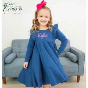 Peut inclure: Robe bleue à manches longues avec une jupe évasée et des épaules à volants. Le nom "Kylie" est brodé en rose sur la poitrine. Un grand nœud rose orne la tête. La robe est de Patsy Ruth Creations.