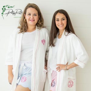 Puede incluir: Dos mujeres vistiendo batas blancas de tejido tipo gofre con ribetes rosas y bordados personalizados. Las mujeres llevan pantalones cortos a juego con ribetes rosas y azules. Las batas tienen un bolsillo en el lado izquierdo. El texto "Patsy Ruth Creations" es visible en la esquina superior izquierda de la imagen.