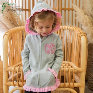 May include: A gray zip-up hoodie featuring a pink gingham ruffle trim along the hood, cuffs, and hem. The garment includes a pink embroidered monogram on the left chest area. The design includes a full-length front zipper and two front pockets.