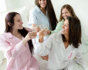 May include: Four individuals wearing button-down pajama sets in light blue, white, and pink pinstripes. The white pajama top features a pink embroidered letter J on the chest pocket. Each person holds a clear glass flute for a toast.