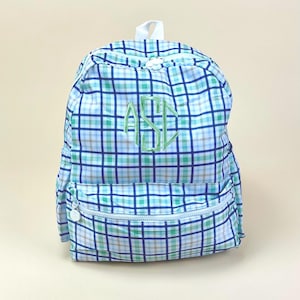 May include: A blue, green, and white plaid backpack with a front zipper pocket and a top handle. The backpack features a green embroidered monogram on the front. The bag has a white zipper and a white zipper pull.