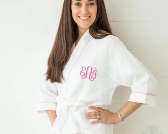 May include: A white waffle-weave robe featuring three-quarter length sleeves, a tie waist, and two front pockets. The robe is accented with pink piping along the edges and displays a pink embroidered monogram on the left chest area.