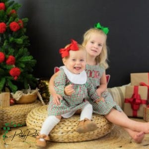 Puede incluir: Dos niños pequeños con vestidos a cuadros verdes y rojos a juego con cuellos blancos y lazos en el pelo. Uno lleva un lazo rojo, el otro un lazo verde. Están sentados en un otomán tejido cerca de un árbol de Navidad.