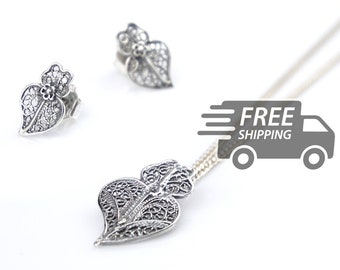 May include: A silver jewelry set consisting of a necklace with a heart-shaped filigree pendant and a pair of matching heart-shaped filigree stud earrings. Each piece features intricate metal wirework patterns and a small flower detail.