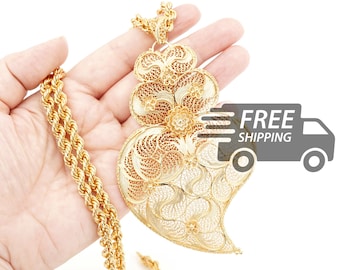 May include: A gold-colored necklace featuring a large, heart-shaped pendant with intricate filigree wirework patterns and a thick rope-style chain. The pendant displays a detailed openwork design with small floral accents throughout the metal surface.
