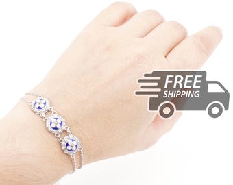 Pode incluir: Uma pulseira de tom prateado com três elos circulares, cada um contendo um centro de esmalte azul profundo com um design de flor branca de quatro pétalas, rodeado por uma borda de pequenas pedras claras numa corrente dupla fina.