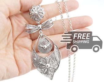 May include: A silver filigree pendant necklace featuring a flower-shaped top, a bow-shaped middle section, and a large teardrop-shaped bottom piece containing a smaller flower design. The pendant hangs from a delicate silver chain.