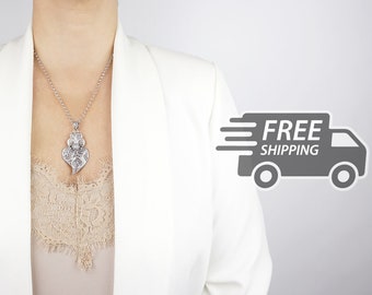 May include: A silver-toned heart-shaped pendant necklace featuring intricate filigree metalwork and a central floral motif, suspended from a delicate silver chain, worn over a beige lace-trimmed top and a white blazer.