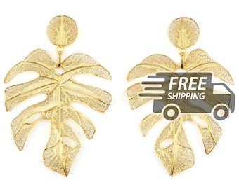 May include: A pair of gold-toned dangle earrings featuring a circular stud connected to a large, intricate monstera leaf pendant. The leaf design is crafted with a delicate, openwork filigree pattern that mimics the veins of a plant.
