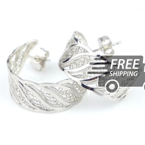 Puede incluir: Un par de pendientes de aro de filigrana plateada con un diseño ondulado. Los pendientes se muestran sobre un fondo blanco.