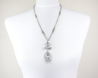 May include: A silver-toned necklace featuring a delicate chain with elongated filigree links. The pendant consists of a small floral element, a bow-shaped detail, and a larger teardrop-shaped frame containing a floral motif and clear crystals.
