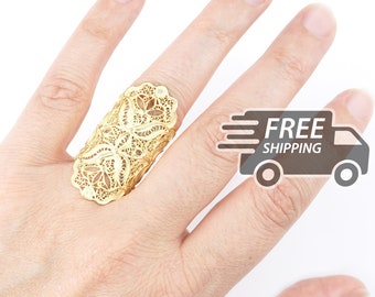 May include: A gold-toned ring featuring an elongated, intricate filigree design with delicate, lace-like metalwork patterns. The ring covers a significant portion of the finger, showcasing a detailed, openwork structure with symmetrical floral-inspired motifs.