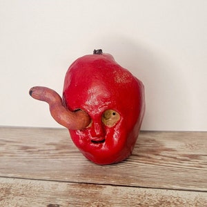 May include: A red ceramic sculpture of a face with a worm for an eye. The sculpture is on a wooden surface.