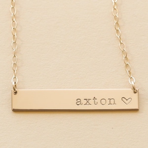 May include: Gold bar necklace with the name "axton" and a small heart engraved on it.