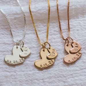 May include: Three delicate necklaces with heart-shaped pendants. Each pendant has a smaller heart charm with initials and a date engraved on the larger heart. The necklaces are silver, gold, and rose gold.